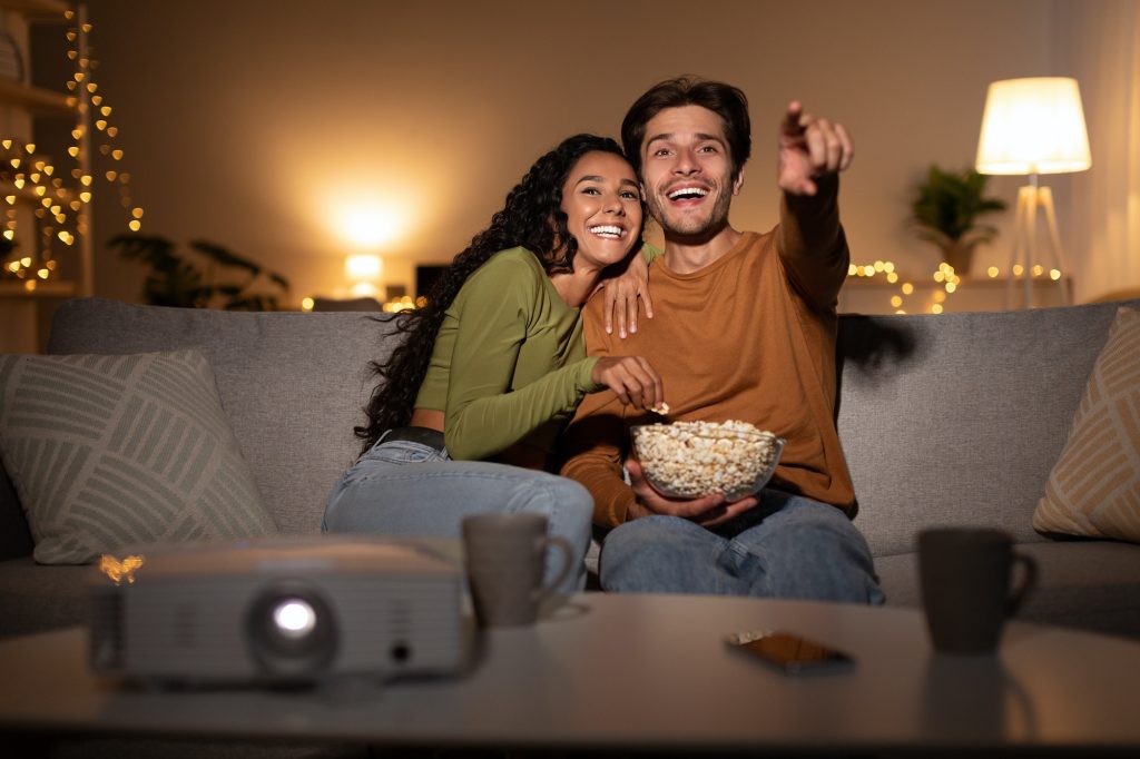 Joyful Couple Watching Film Online Using Home Cinema Projector Indoors